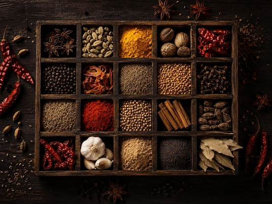 A dark wooden spice box with 16 compartments filled with various whole and ground spices, herbs, and seasonings, arranged on a dark rustic wooden surface with scattered red chilies and star anise.