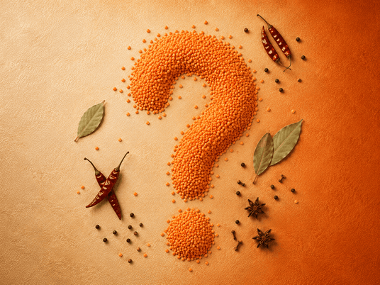 A creative flat-lay composition of orange lentils shaped into a large question mark with a smaller pile below, surrounded by whole spices, star anise, bay leaves, and dried red chilies on a textured orange background.
